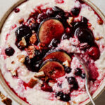 An image of Easy Barley Porridge topped mixed berries, figs and pecans. The porridge is in a grey bowl with a spoon in the middle. The bowl is sitting on a white backdrop with pecans sprinkled around.