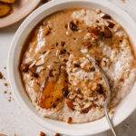 Perfect Steel Cut Oatmeal topped with almond butter, almonds, maple syrup, cinnamon and caramelized bananas. The oatmeal is in a white bowl with a spoon in the middle. The bowl is sitting on a white backdrop with a plate of caramelized bananas in the top left, a bow of cinnamon in the bottom left, sprinkles of cinnamon and almonds.