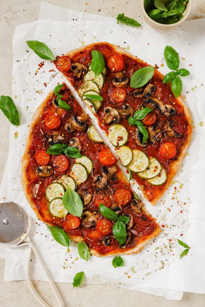 Vegan pizza cooked and coated with homemade marinara sauce and topped with sliced veggies. The pizza is placed on parchment paper and sprinkled with basil. The parchment paper is sitting on a white backdrop with a pizza cutter in the bottom left and a bowl of fresh basil in the top right.