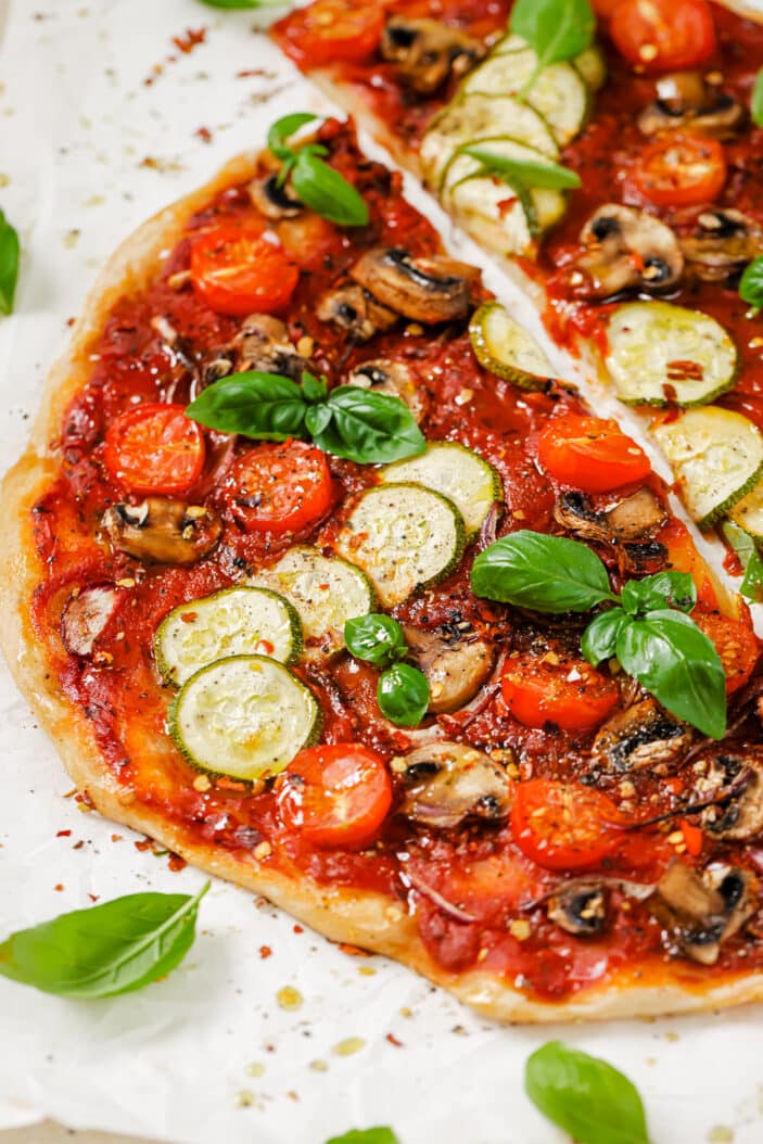 Vegan pizza cooked and coated with homemade marinara sauce and topped with sliced veggies. The pizza is placed on parchment paper and sprinkled with basil. The parchment paper is sitting on a white backdrop.