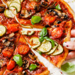 Side angle image of vegan pizza decorated with vegetables cut into slices and a hand is grabbing a slice and pulling it away. The pizza is sitting on parchment paper.