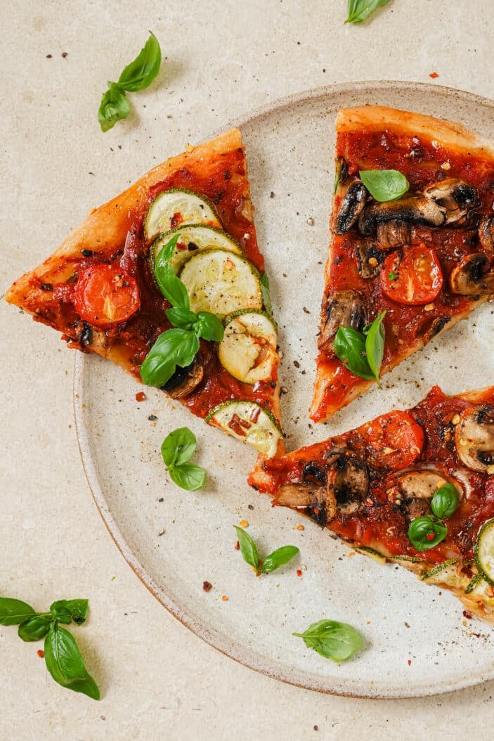 three slices of vegan pizza topped with sliced veggies. The pizza is on a white plate. The plate is sitting on a white backdrop with fresh basil sprinkled around.
