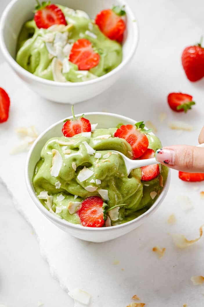 matcha nice cream in a white bowl with coconut flakes and strawberries on top. A spoon is scooping some of the matcha nice cream out of the bowl.