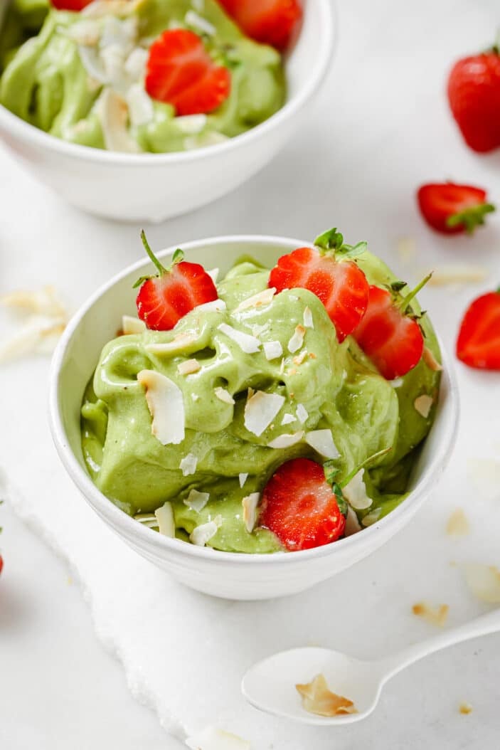 matcha nice cream in two white bowls, sprinkled with coconut flakes and topped with fresh strawberries on a white backdrop