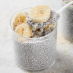 coconut chia pudding in a glass cup topped with banana and the toasted coconut. A spoon is scooping out some of the chia pudding.