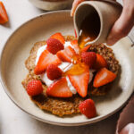 whole wheat pancakes on a white plate topped with coconut yogurt, cut strawberries and raspberries. There is a hand pouring maple syrup onto the pancakes in a small brown jug. The pancakes are on a pinky brown countertop with a bowl of strawberries in the background.