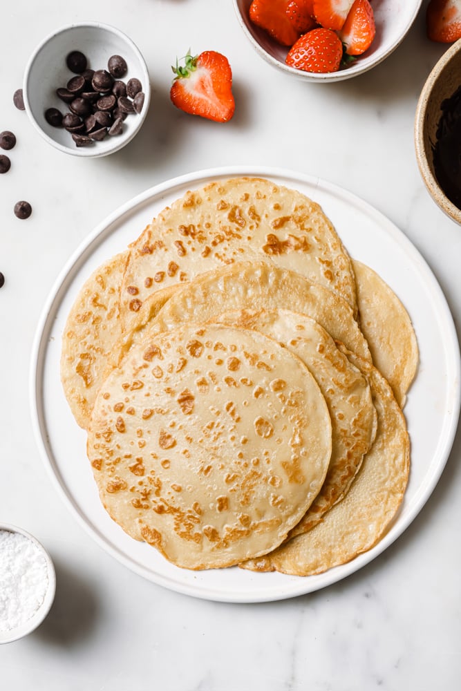 Cooked crepes placed on a white bowl sitting on a white backdrop. There are two small bowls in the top left and right hand corner, one is filled with chocolate and the other is filled with strawberries.