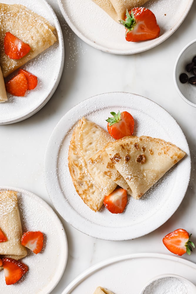 Top down image of a few plates of crepes. There are two crepes on each plate and they are ready to be decorated. Sliced strawberries have been placed on the plates as well. The plates are sitting on a white backdrop.