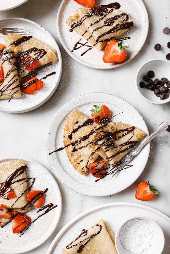 Top down image of four plates of crepes. Each white plate holds two crepes that have been drizzled with chocolate and sprinkled with sliced strawberries. The white plates are on a white backdrop. 