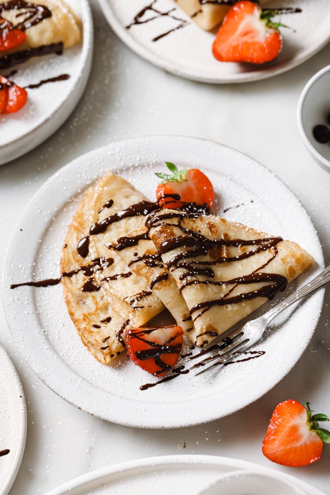 Image of two crepes on a white plate that have been drizzled with chocolate and sprinkled with sliced strawberries. The white plate is on a white backdrop.  There is a strawberries in the lower right hand corner of the frame.