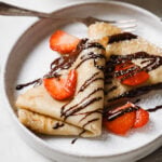 Image of two crepes on a white plate that have been drizzled with chocolate and sprinkled with sliced strawberries. The white plate is on a white backdrop.