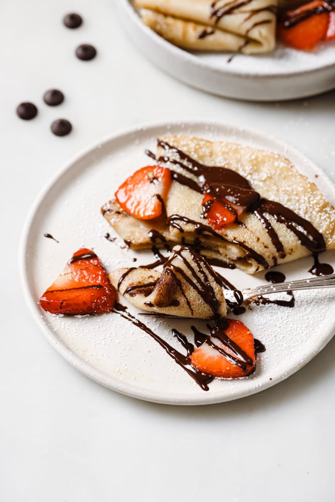 A crepe that is on a white plate and drizzled with melted chocolate and sprinkled with strawberries. The white plate is on a white backdrop.