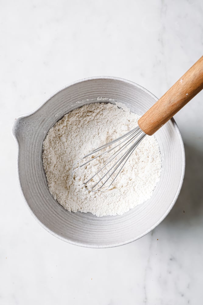 Flour and salt in a large mixing bowl with a whisk in the middle that's been used for mixing. The white bowl is on a white backdrop. 
