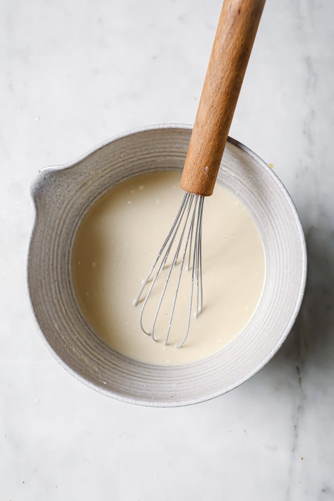 Crepe ingredients that have been whisked together with a whisk to make a smooth batter. The batter is in a white mixing bowl on a white backdrop.