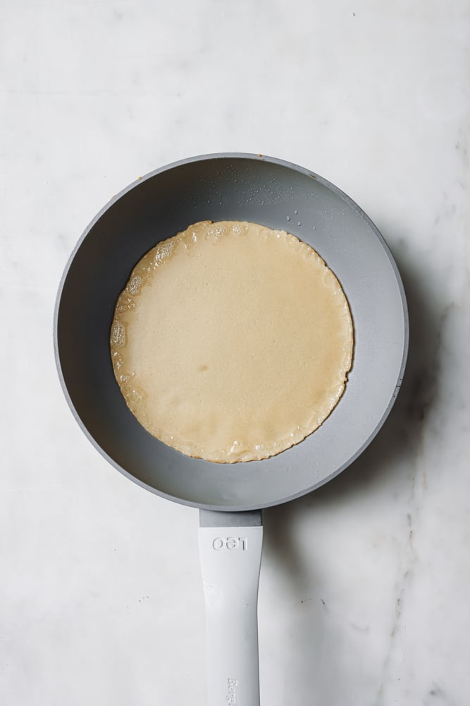 Crepe batter that's been swirled on a pan to make a circle shape. The pan is grey and it's sitting on a white backdrop.