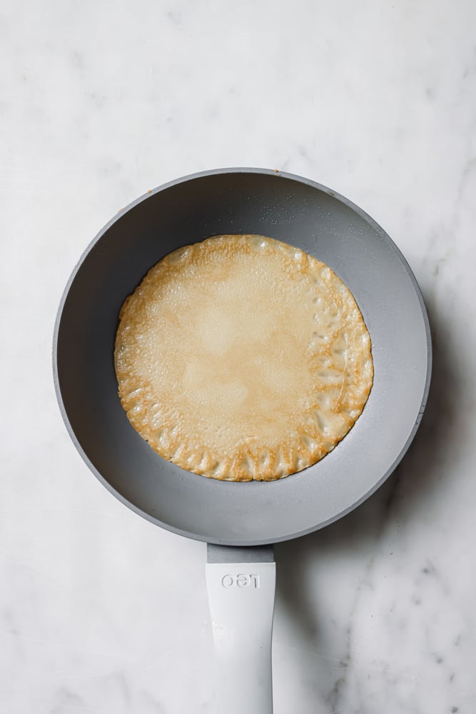 Cooked crepe in a skillet. The skillet is grey and it's sitting on a white backdrop.