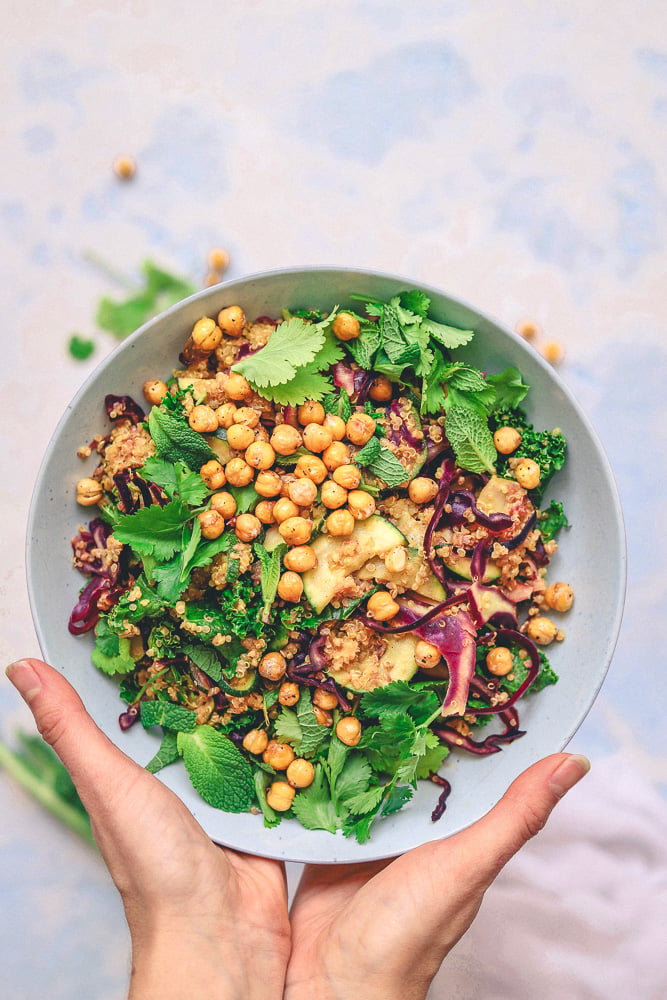 A person holds a bowl of salad with roasted chickpeas, greens, herbs, cucumber slices, and a purple vegetable on a light background.
