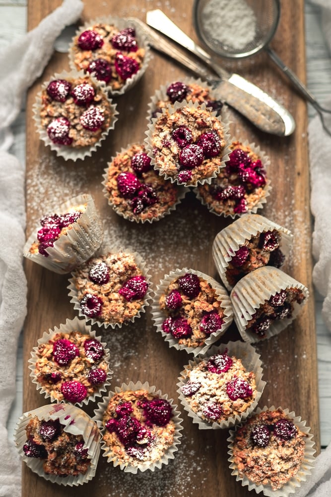 A flatlay image of healthy raspberry oat muffins. The muffins are on a wooden cutting board with a knife and a sieve full of icing sugar in the top right corner and a sprinkle of icing sugar on the muffins. The cutting board is sitting on a white  cloth.