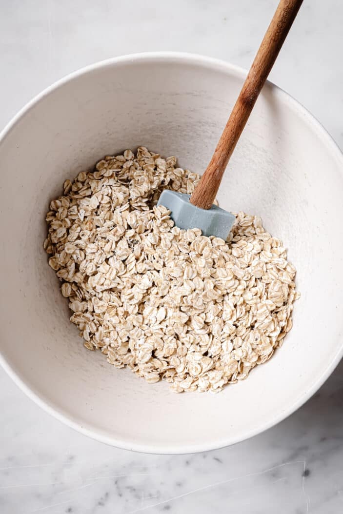 The oats and chia seeds used to make the healthy raspberry oat muffins. They are mixed together in a large white bowl with a spatula in the middle. The bowl is sitting on a white backdrop.