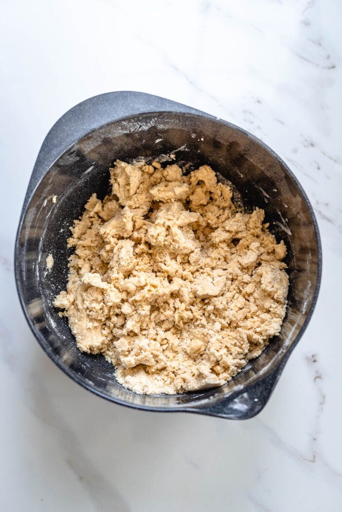 All of the ingredients to make vegan short bread cookies combined to form a patchy dough. This mixture is in a grey bowl. The bowl is sitting on a white backdrop.