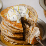 Lemon poppy seed pancakes, stacked, and topped with a dollop of coconut yogurt, lemon zest and maple syrup. The pancakes are stacked on a grey plate with a slice taken out to show the fluffy inside. The plate is sitting on a white backdrop with lemon zest in the bottom left, lemon slices in the bottom right.