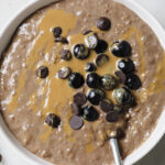A bowl of chocolate oatmeal topped with blueberries, chocolate chips and drizzled with peanut butter. The chocolate oatmeal is in a white bowl on a white backdrop.