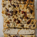 Vegan focaccia bread topped with sun dried tomatoes, red onion, olives and rosemary. The bread is cut into slices, with one flipped to show the inside texture. The focaccia is placed on a white backdrop.