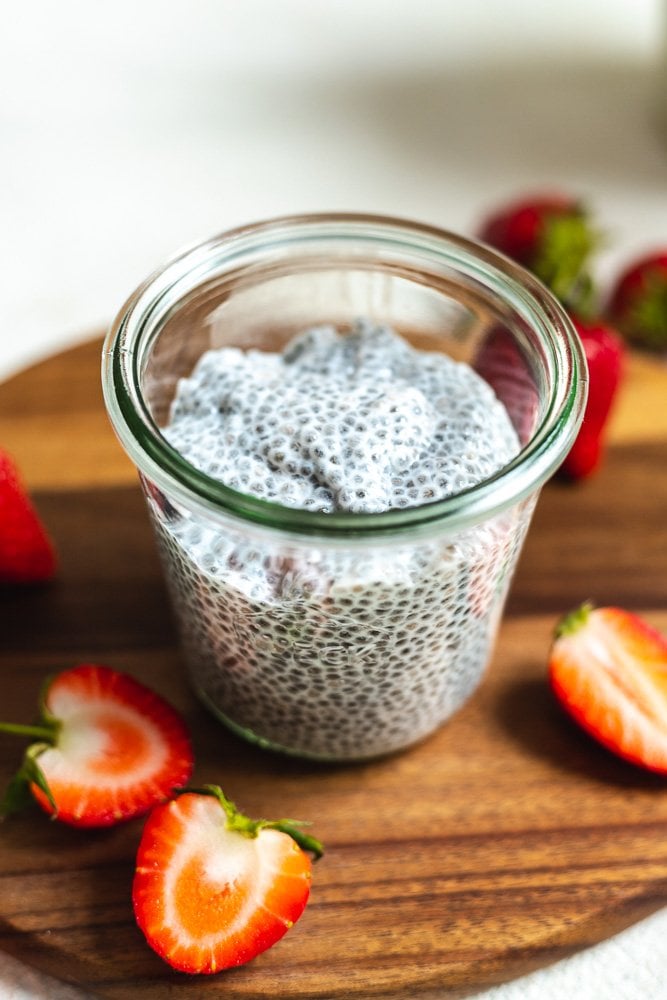 Easy 3-Ingredient Chia Pudding placed in a glass  jar. The jar is sitting on a wooden cutting board with sliced strawberries. The backdrop is white.