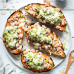 Four healthy sweet potato skins loaded with white bean, guacamole and are drizzled with a citrus tahini dressing. The sweet potato skins are on a white plate. The white plate is sitting on a white backdrop with a bowl of sesame seeds in the top right, a wooden spoon in the bottom right, lavender in the top left and a tea towel in the bottom left.