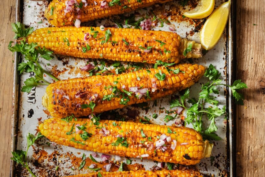 oven-roasted corn on the cob that's been broiled until lightly charred. Sprinkled with fresh herbs, red onion, salt and pepper. The corn is still on the baking tray on a wood backdrop