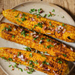 image of oven roasted corn on the cob slathered in miso butter and sprinkled with fresh herbs, red onion, salt and pepper. The corn is on a beige plate on a wood backdrop.