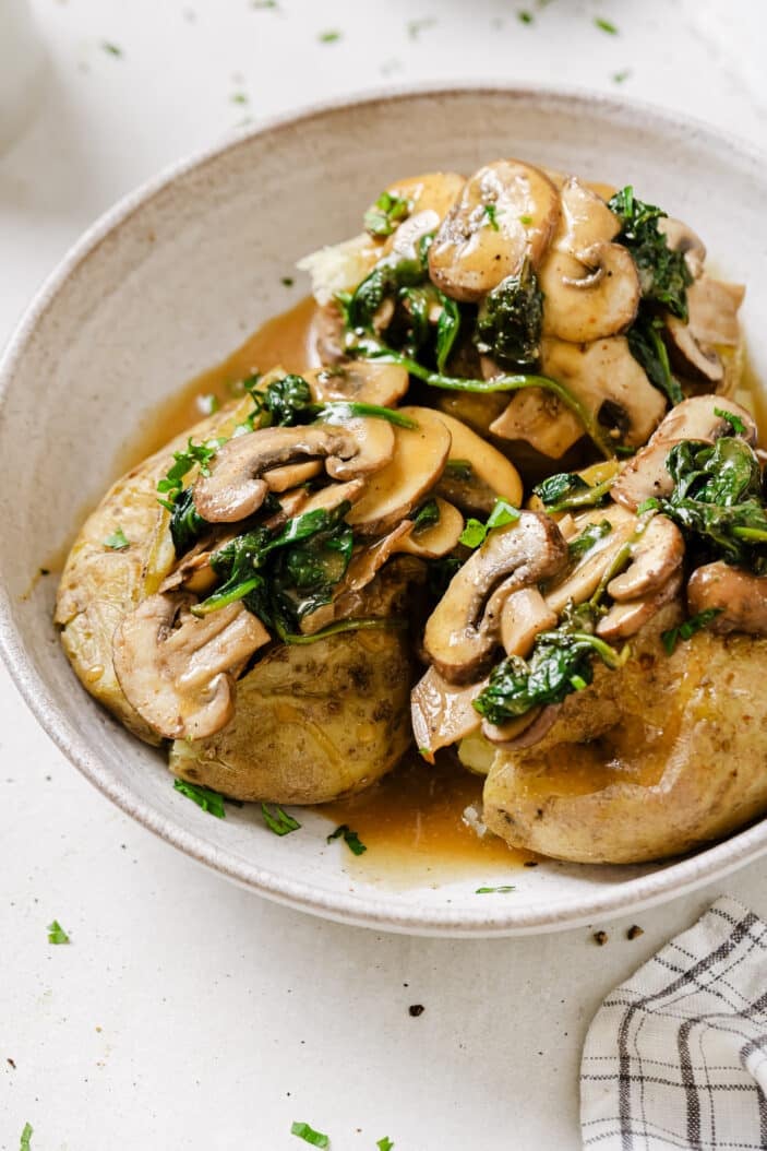 A white bowl of baked potatoes stuffed with mushroom, spinach and gravy on a white backdrop.