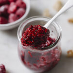 Raspberry chia jam in a small glass jar with a silver spoon scooping out some of the chia jam. In the back right hand corner is a white bowl filled with raspberries. The jar of chia jam is sitting on a white backdrop.