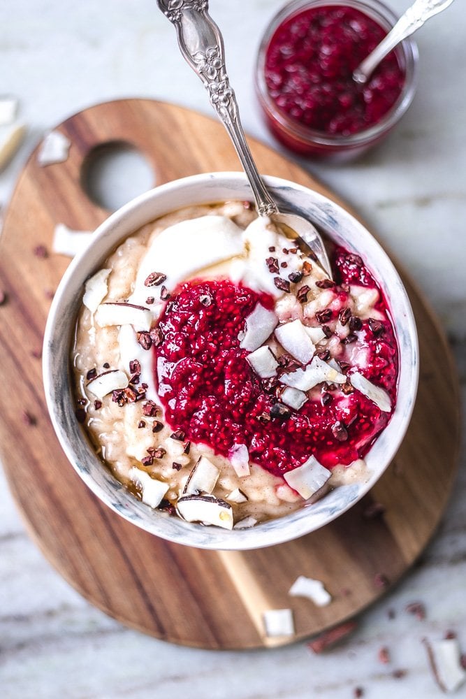 image of raspberry chia jam swirled into a bowl of rice pudding. The bowl is sitting on a wood cutting board on a white backdrop.