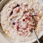 Vegan Rice Pudding dolloped with mixed berry compote and sprinkled with spices. The pudding is scooped into a white bowl with a spoon in the middle. The bowls are sitting on a white backdrop that was sprinkled with spices, a cup of maple syrup and spoons placed in the bottom right.
