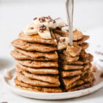 stacked pancakes with a corner cut out and a fork wedges into all the layers of the pancake. The pancakes look fluffy and light. They are topped with bananas and cacao nibs.