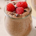 chocolate chia pudding in a clear glass jar topped with raspberries. The glass is sitting on a brown countertop with sprinkled of chocolate on the countertop.