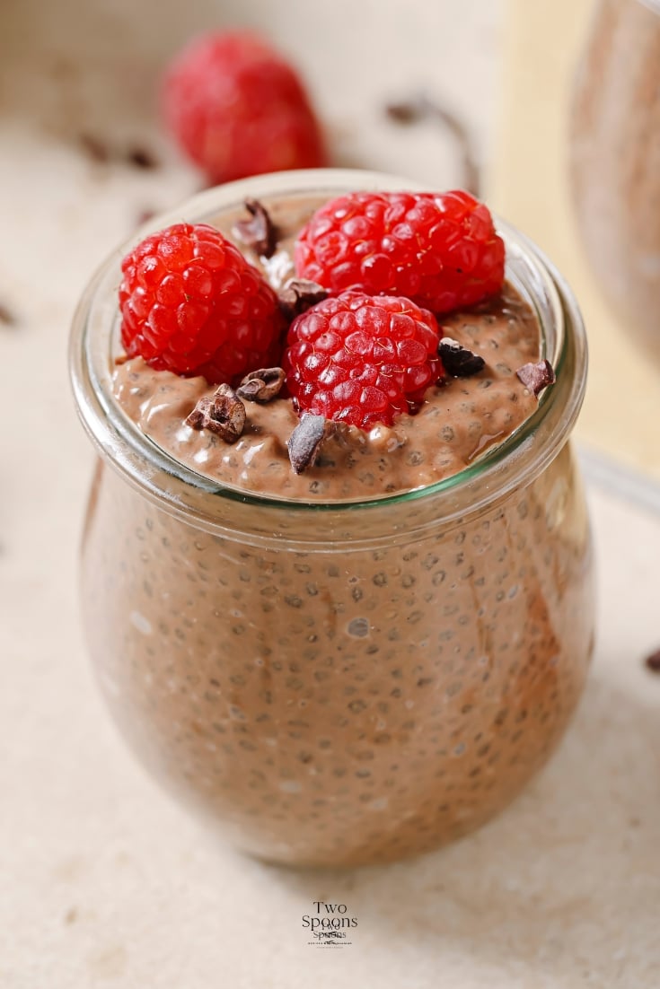 chocolate chia pudding in a clear glass jar topped with raspberries. The glass is sitting on a brown countertop with sprinkled of chocolate on the countertop.