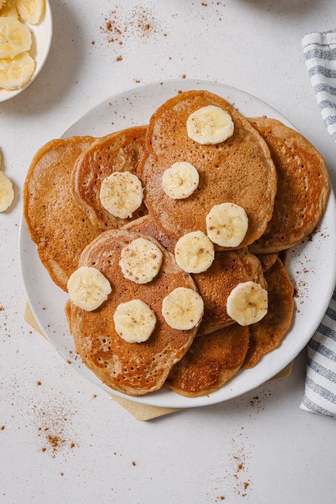 A stack of fluffy vegan banana pancakes topped with bananas. The pancakes are stacked on a white plate. The plate is sitting on a white backdrop with a plate of sliced bananas in the top left corner.