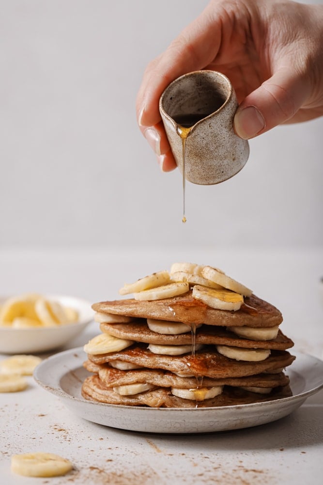 A stack of vegan banana pancakes with banana slices throughout the layers. The pancakes are stacked on a white plate with a cup of maple syrup being poured on top. The plate is sitting on a white backdrop with a plate of banana slices in the top left.