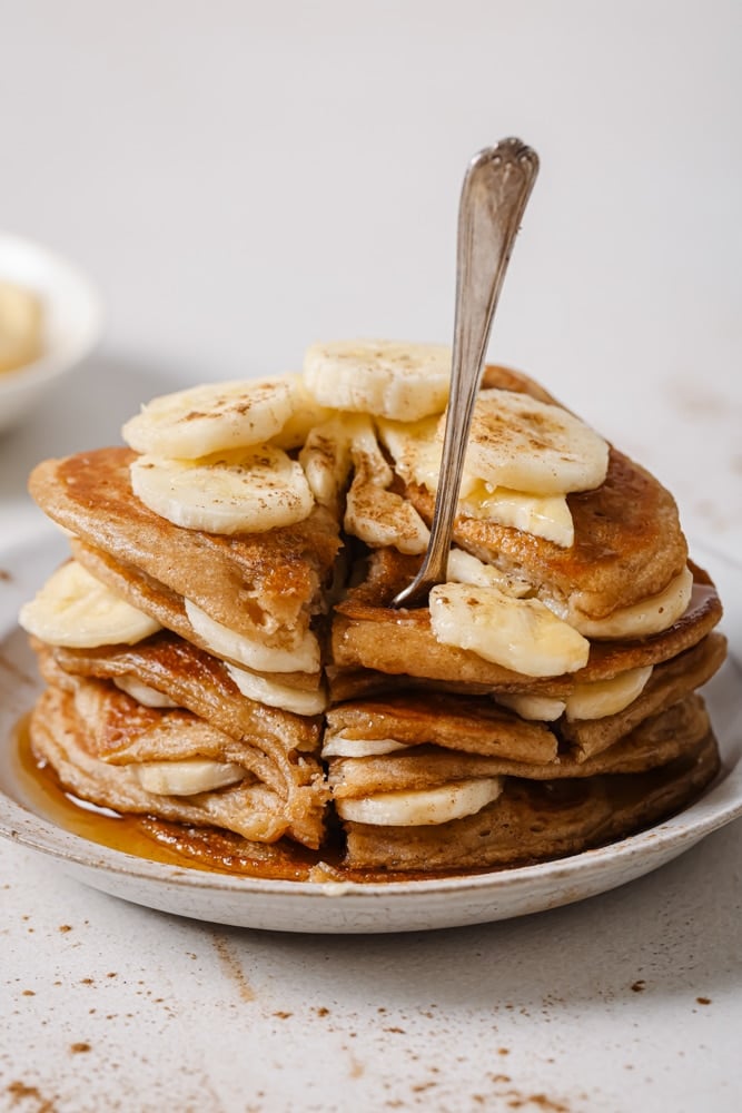 A stack of fluffy vegan banana pancakes, the stack consists of pancakes and sliced bananas. The pancakes are stacked on a white plate with a fork holding a slice of the pancakes. The plate is sitting on a white backdrop.