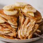 A stack of fluffy vegan banana pancakes, the stack consists of pancakes and sliced bananas. The pancakes are stacked on a white plate with a fork holding a slice of the pancakes, to show the fluffy inside. The plate is sitting on a white backdrop.