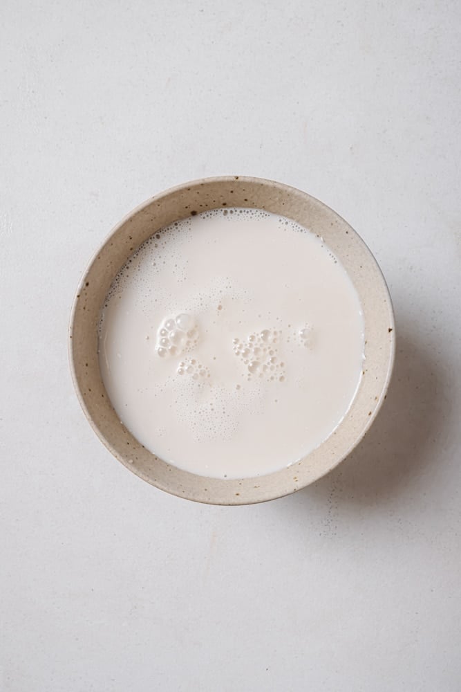Plant-based milk combined with apple cider vinegar to make a vegan buttermilk. The buttermilk is poured into a grey bowl. The bowl is sitting on a white backdrop.