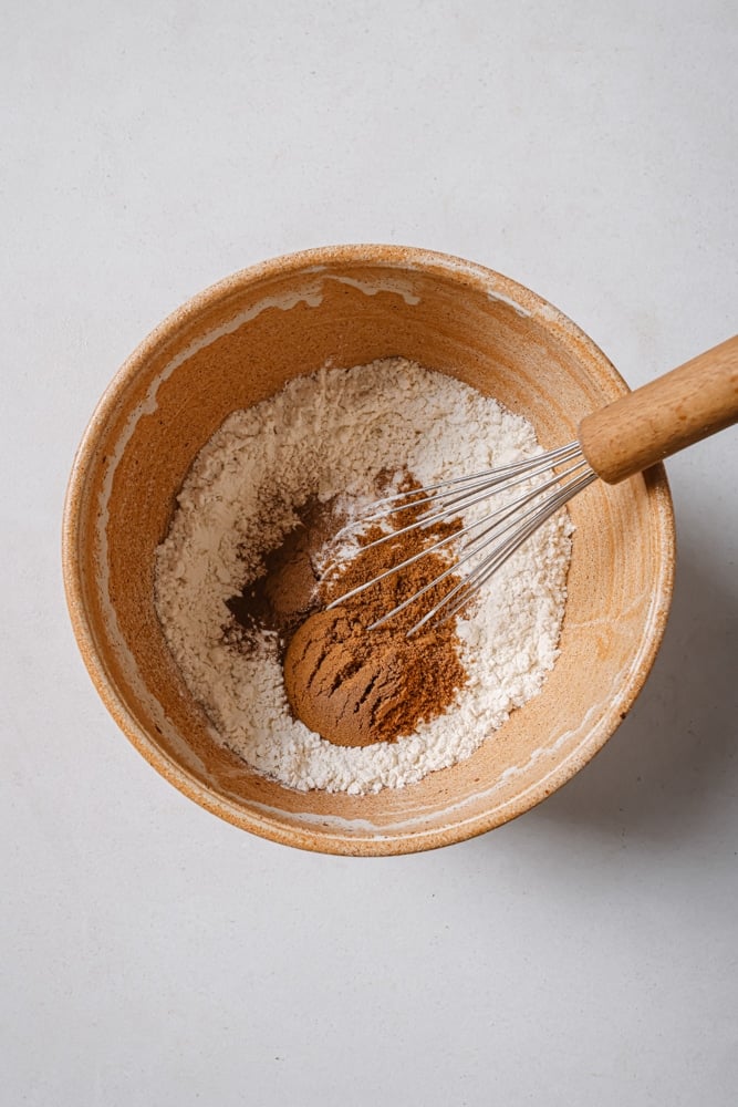 All of the dry ingredients to make the fluffy vegan pancakes placed into a brown bowl. The bowl is sitting on a white backdrop with a wooden whisk inside the bowl.