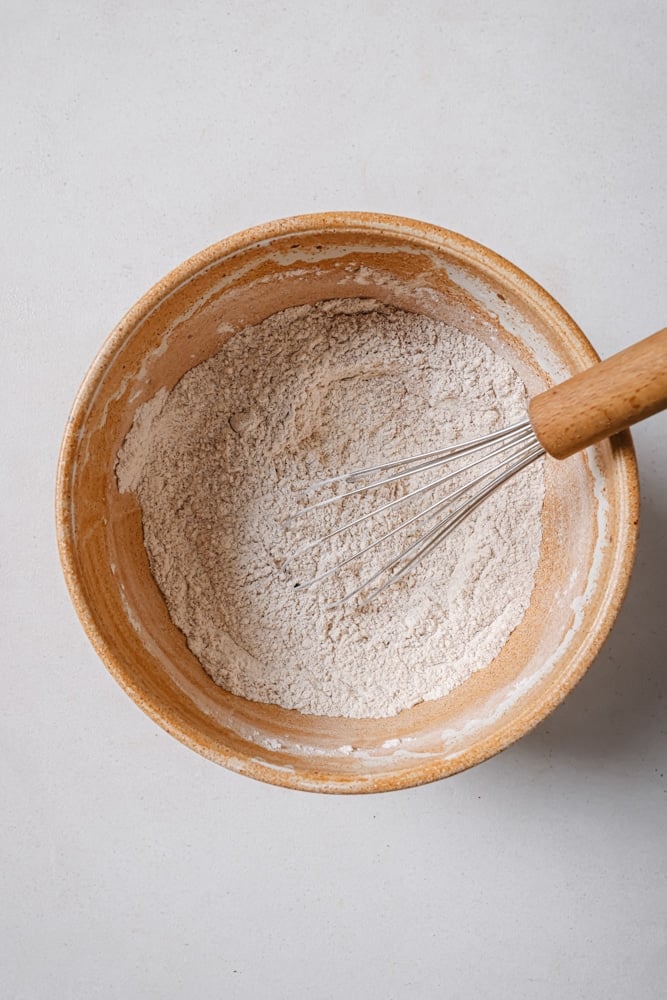 All of the dry ingredients to make the fluffy vegan pancakes whisked together. The flour mixture is in a brown bowl with a wooden whisk in the middle. The bowl is sitting on a white backdrop.