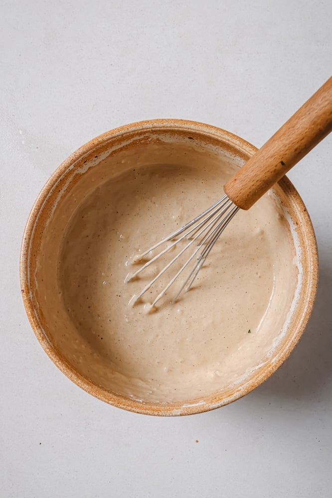 All of the ingredients to make the fluffy vegan banana pancakes combined to make a batter. The batter is in a brown bowl with a whisk in the middle. The bowl is sitting on a white backdrop.