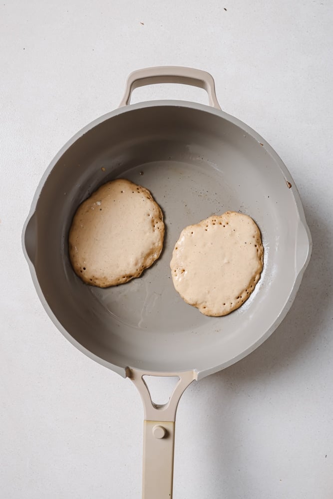 The fluffy vegan pancake batter poured into a grey skillet. The batter is being cooked on the bottom with bubbles forming. The skillet is sitting on a white backdrop.