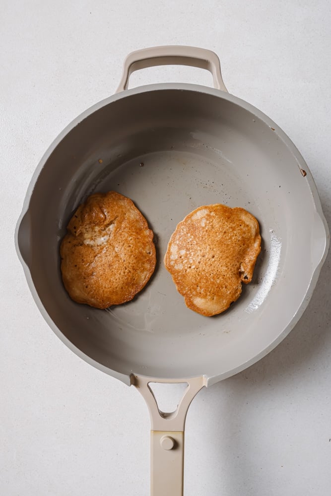 The fluffy vegan banana pancakes cooked on a grey skillet. The skillet is sitting on a white backdrop.
