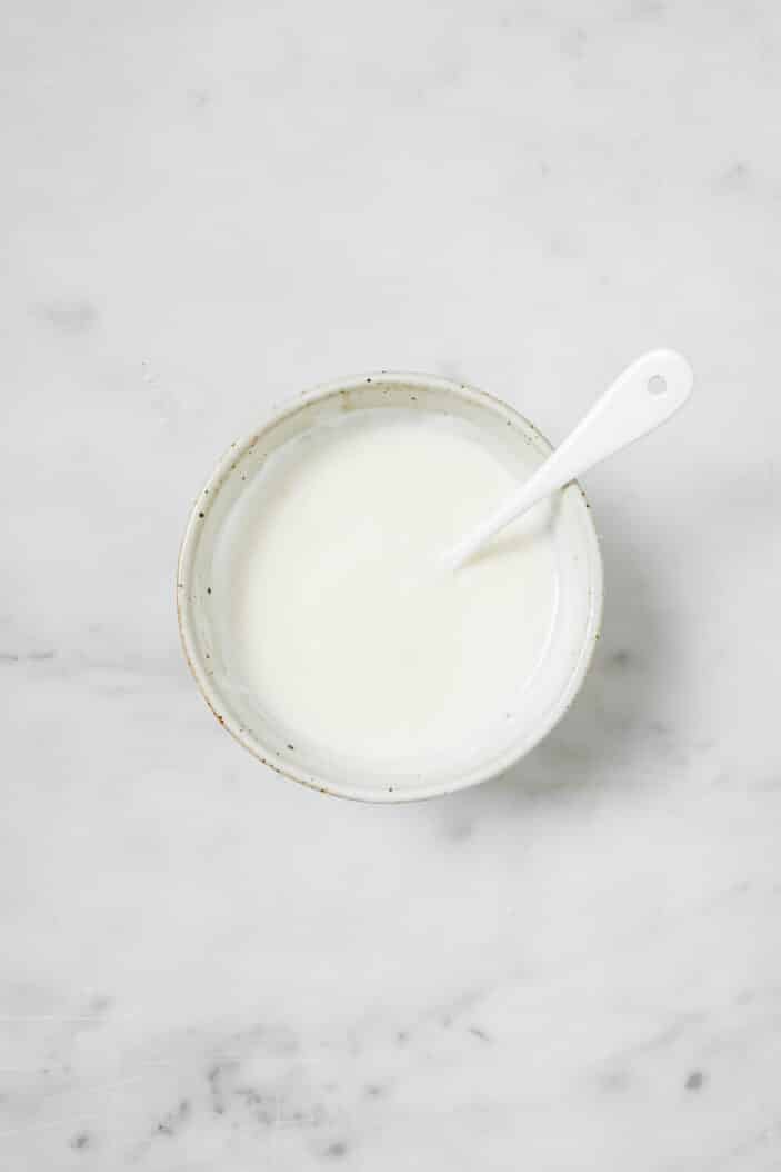 simple vegan sour cream using coconut yogurt, salt and lime juice that's been stirred together with a spoon. The bowl is sitting on a white marble backdrop.