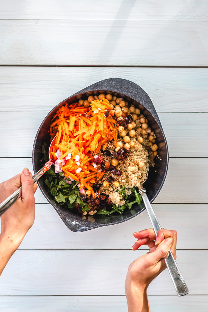 A person mixes grated carrots, chickpeas, quinoa, greens, onions, and raisins in a black bowl with two utensils on a white wooden table.